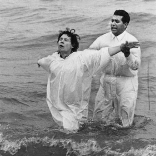 A man wearing a white shirt and pants is holding the arms of a woman who is wearing a white robe and spreading her arms out. They are both standing knee deep in water. Caption reads: 'The Rev. Ray E. Lopez, pastor of the Apostolic Assembly of the Faith in Christ Jesus, 606 S. 5th St., baptized Mrs. Maria Martinez Saturday in Lake Michigan. Four persons were baptized in the ceremonies at Bradford beach.'                         Un hombre vestido con camisa y pantalones blancos sostiene los brazos de una mujer que lleva una bata blanca y extiende los brazos. Ambos están parados en el agua hasta las rodillas. El título dice: 'El reverendo Ray E. López, pastor de of the Apostolic Assembly of the Faith in Christ Jesus, 606 S. 5th St., bautizó a la Sra. María Martínez el sábado en el lago Michigan. Cuatro personas se bautizaron en las ceremonias de Bradford Beach'.